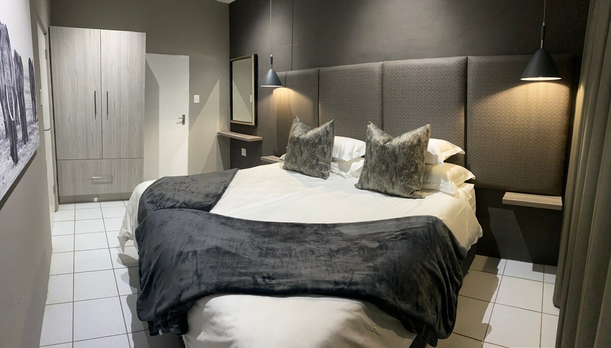 A spacious sleeping area features a large bed with plush pillows and a soft throw blanket. Dark accent walls are complemented by bedside lamps, while a mirror and wardrobe are visible in the room. Natural light enters through a nearby window, enhancing the room's ambiance.