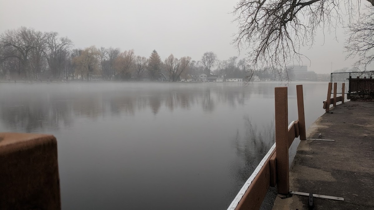 The tranquil water of the Fox River is depicted, reflecting the foggy surroundings. Trees line the bank, and a wooden railing is visible along the edge, providing a sense of connection to the serene landscape. The scene captures a calm, early morning atmosphere.