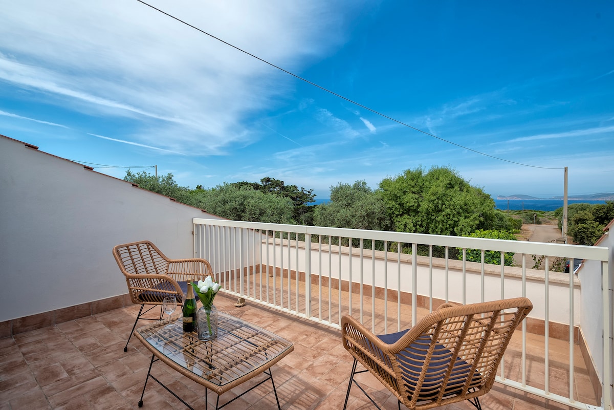 A balcony is showcased with a table and two wicker chairs, offering a view of greenery and the distant sea. The natural blue sky complements the serene outdoor space, creating an inviting area for relaxation.