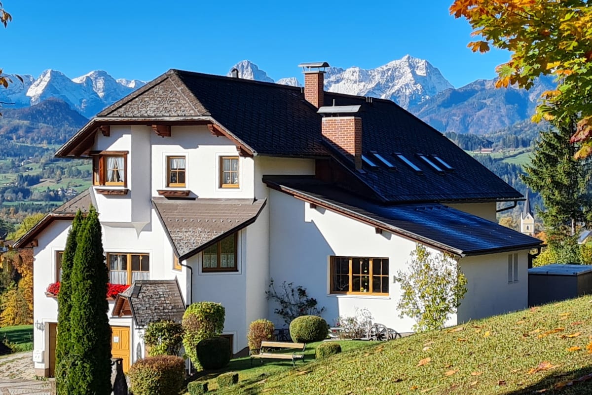 The exterior of a multi-level house is presented, featuring a combination of white walls and wooden accents. Surrounding greenery and well-maintained shrubs frame the structure. Snow-capped mountains are visible in the background against a clear blue sky.
