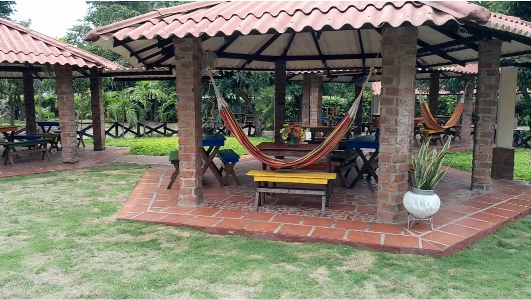 A spacious outdoor area features a covered structure with a tiled floor and brick columns. Colorful hammocks are hung between columns, and tables with benches are arranged around the space. Lush greenery surrounds the area, providing a natural backdrop for relaxation.
