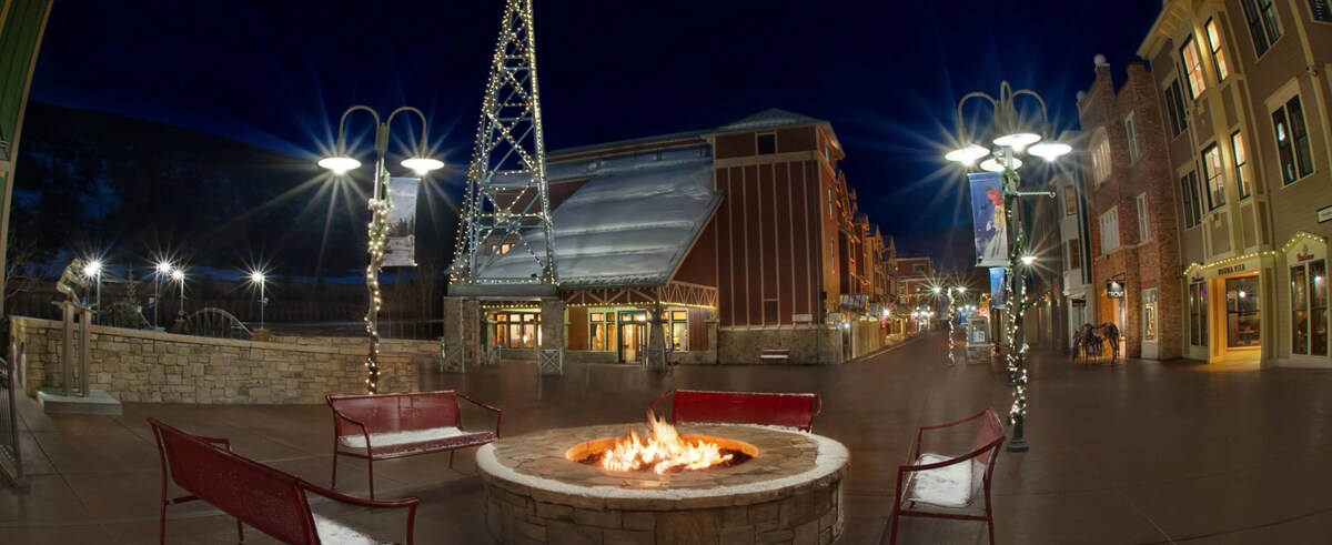 An outdoor plaza features a circular fire pit at its center, surrounded by red benches. The evening scene is illuminated by street lamps and festive lights, while the surrounding buildings showcase a mix of architectural styles. The mountain backdrop adds to the tranquil atmosphere.