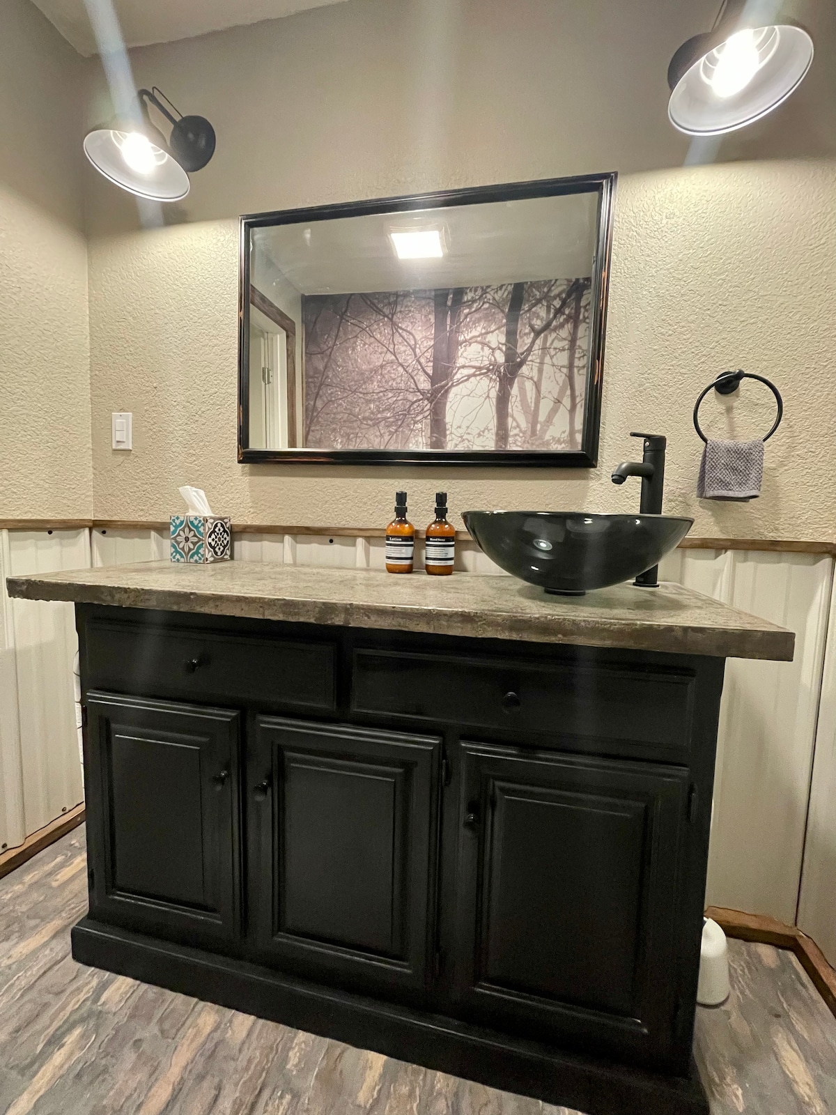 A well-lit bathroom area features a black vessel sink atop a dark countertop, complemented by black cabinetry. A large mirror reflects the space, and soft lighting accentuates the neutral walls and wood detailing. Essential toiletries and a decorative tissue box add functionality.