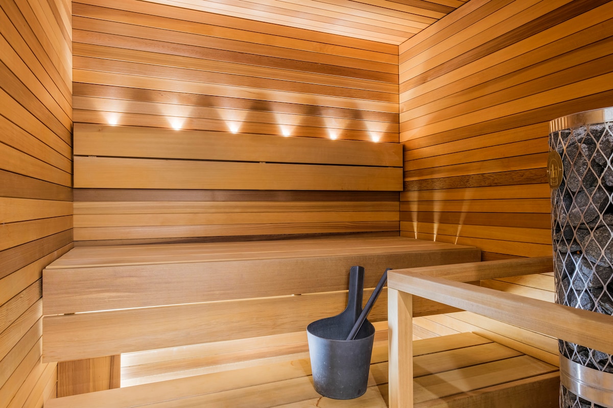 A warm sauna space is showcased, featuring polished wooden walls and benches. Soft recessed lighting is positioned along the wall, illuminating the area. A wooden bucket and ladle are placed beside a stone heater, enhancing the serene atmosphere.