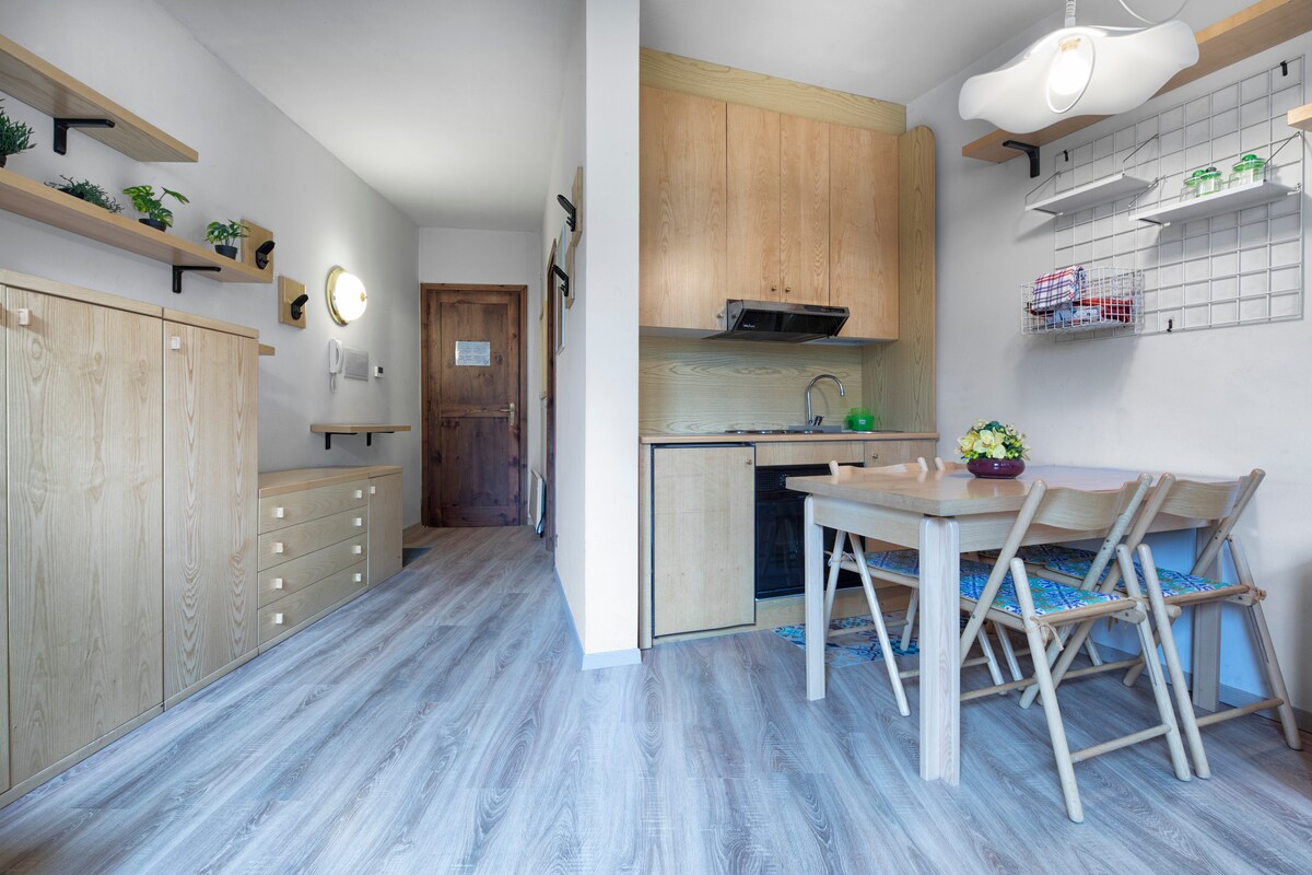 A well-lit dining area is complemented by natural wooden cabinetry and shelving. A round table with four chairs is positioned next to a compact kitchen, featuring modern appliances and decorative plants. The light wood flooring enhances the open and airy feel of the space.