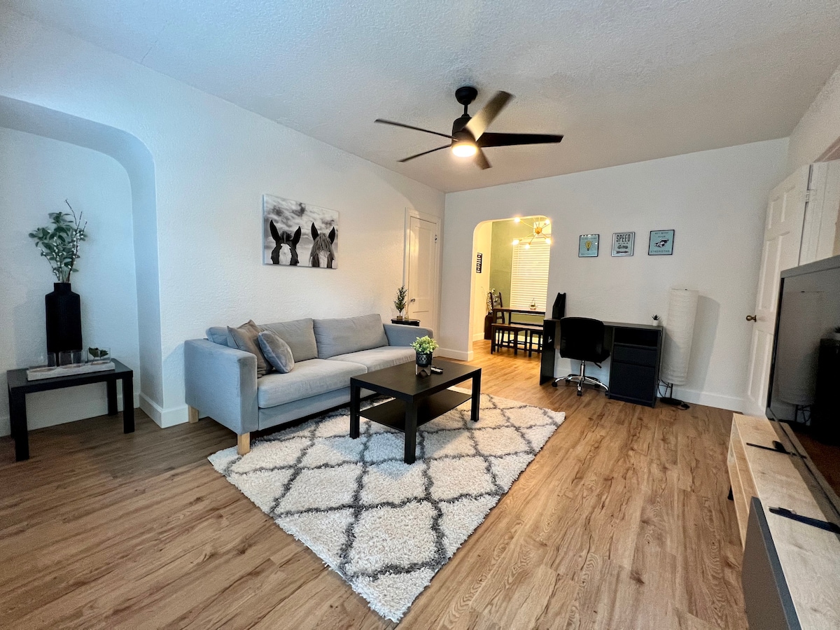 A cozy living area features a soft gray sofa centered on a patterned area rug, complemented by a black coffee table. A ceiling fan and natural light enhance the space, while a small desk and dining area are visible in the background.