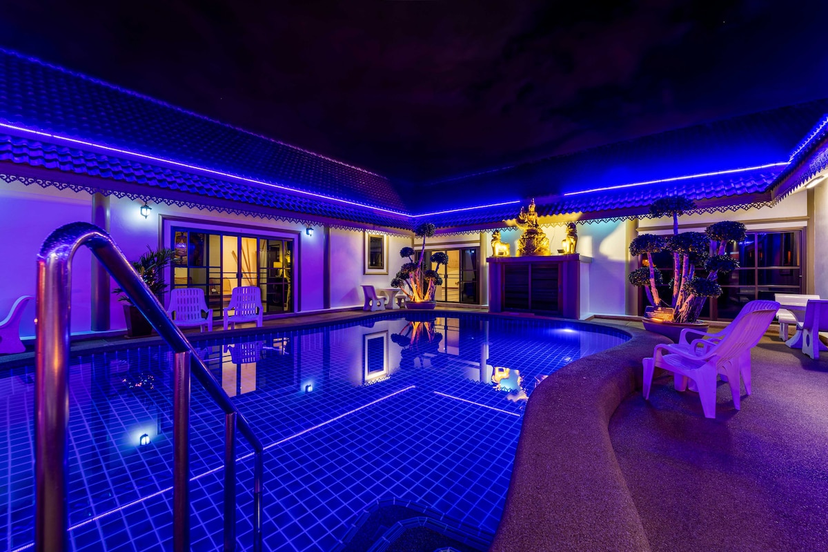 A serene pool area is surrounded by a decorated patio, with multicolored lighting casting a warm glow on the water's surface. Lounge chairs are positioned beside the pool, inviting relaxation. The spacious layout includes tropical plants and a decorative feature nearby, enhancing the tranquil environment.