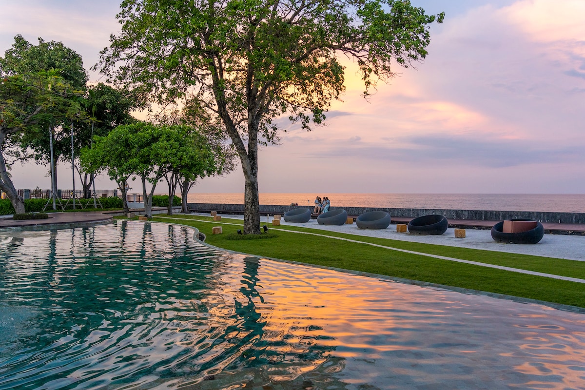 A serene beachfront pool area is surrounded by lush greenery and contemporary seating. The water reflects the soft hues of the sunset, creating a tranquil atmosphere. Comfortable circular lounge chairs are positioned near the pool, offering views of the ocean in the background.