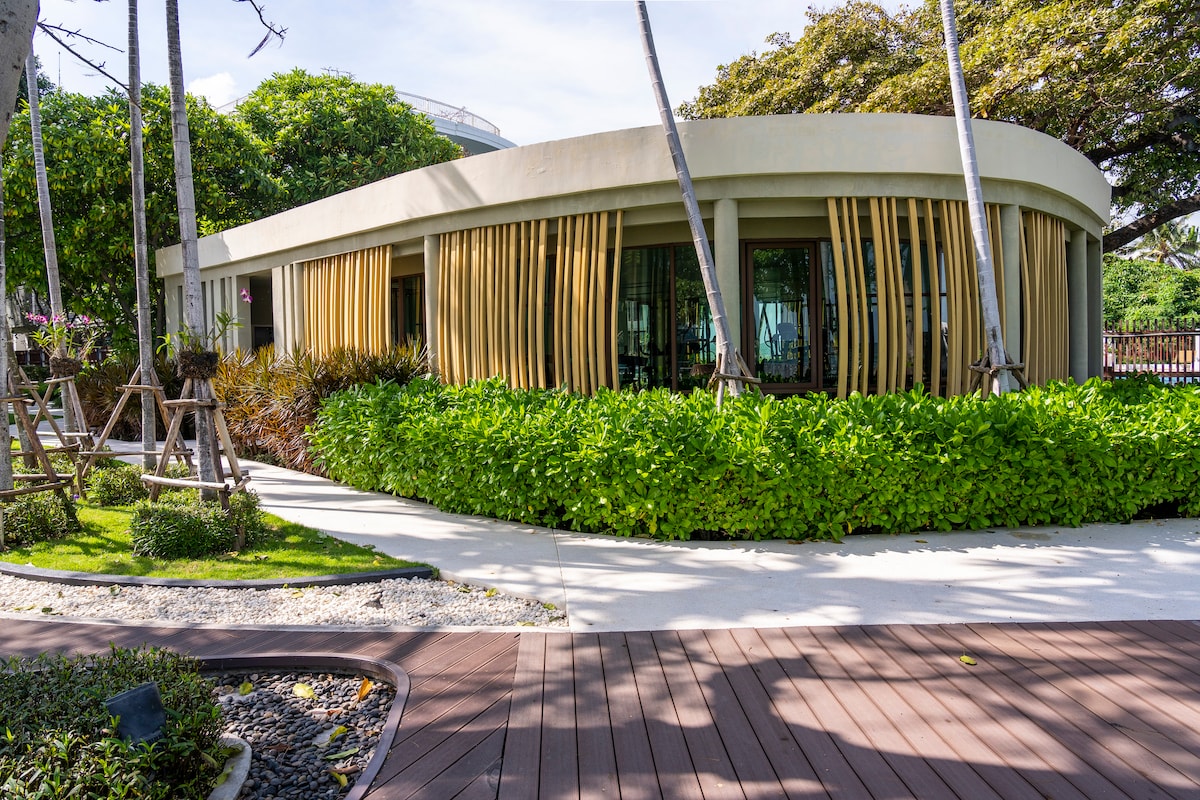 A modern beachfront condo is showcased with curved architectural lines. Lush greenery surrounds the building, with tall palm trees adding to the tropical feel. The exterior features large windows that invite natural light, providing a connection to the outdoor landscaping.