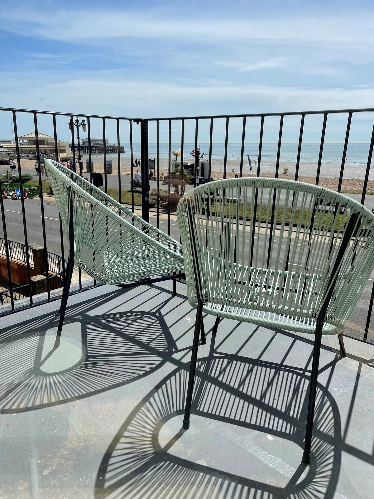 Two stylish chairs are situated on an accessible balcony, with a view of the sea and nearby beach. Natural light creates intriguing shadows across the floor, enhancing the peaceful setting. The balcony railing offers safety while allowing for unobstructed views of the surroundings.