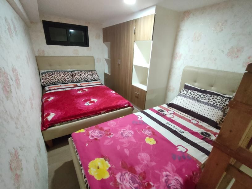 A cozy sleeping area features two spacious beds dressed in patterned bedding. Soft, warm tones are complemented by floral and striped designs. A small closet is visible, along with a window allowing natural light to brighten the room.