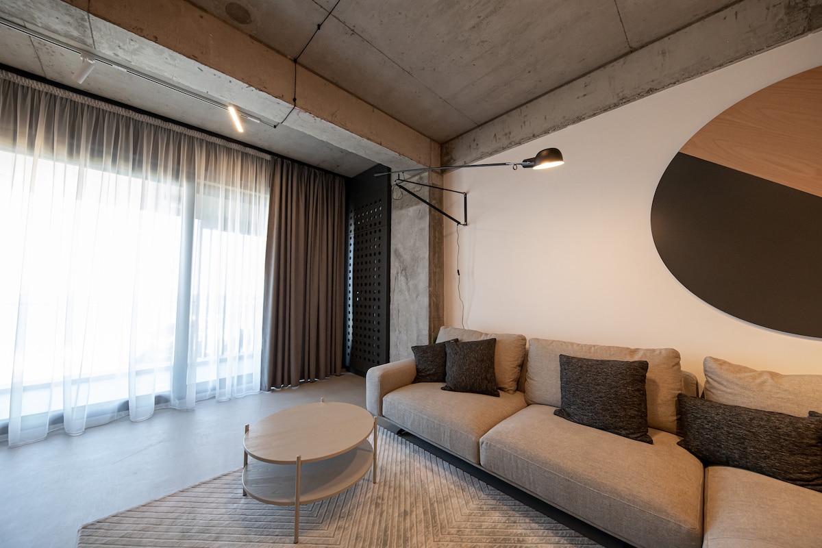 A modern living area features a spacious sofa with decorative cushions, positioned near large glass doors that allow natural light. A round coffee table is centered on a textured rug. Floor-to-ceiling curtains frame the view, adding an element of softness to the industrial design.