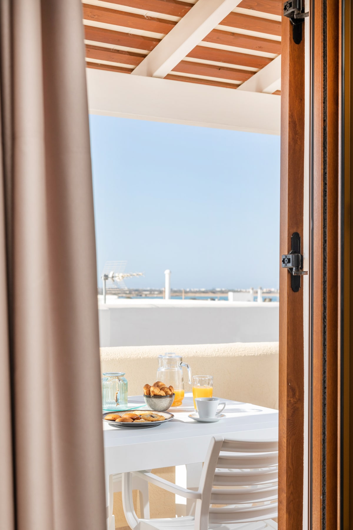 A light-filled terrace is visible through partially open doors, featuring a table set for breakfast. A glass carafe and cups accompany a plate of pastries, while the distant view of the sea can be seen against a clear blue sky.