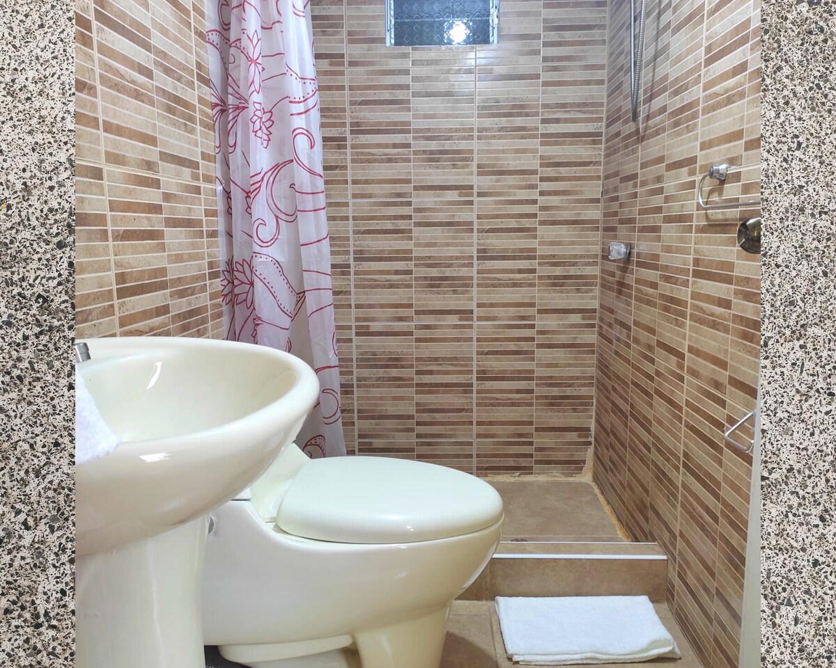 A modern bathroom is shown, featuring warm-toned tiles and a walk-in shower with a clear glass partition. A white shower curtain with a floral design hangs beside a toilet. A cotton bath mat is placed on the floor, enhancing the bathroom's functionality.