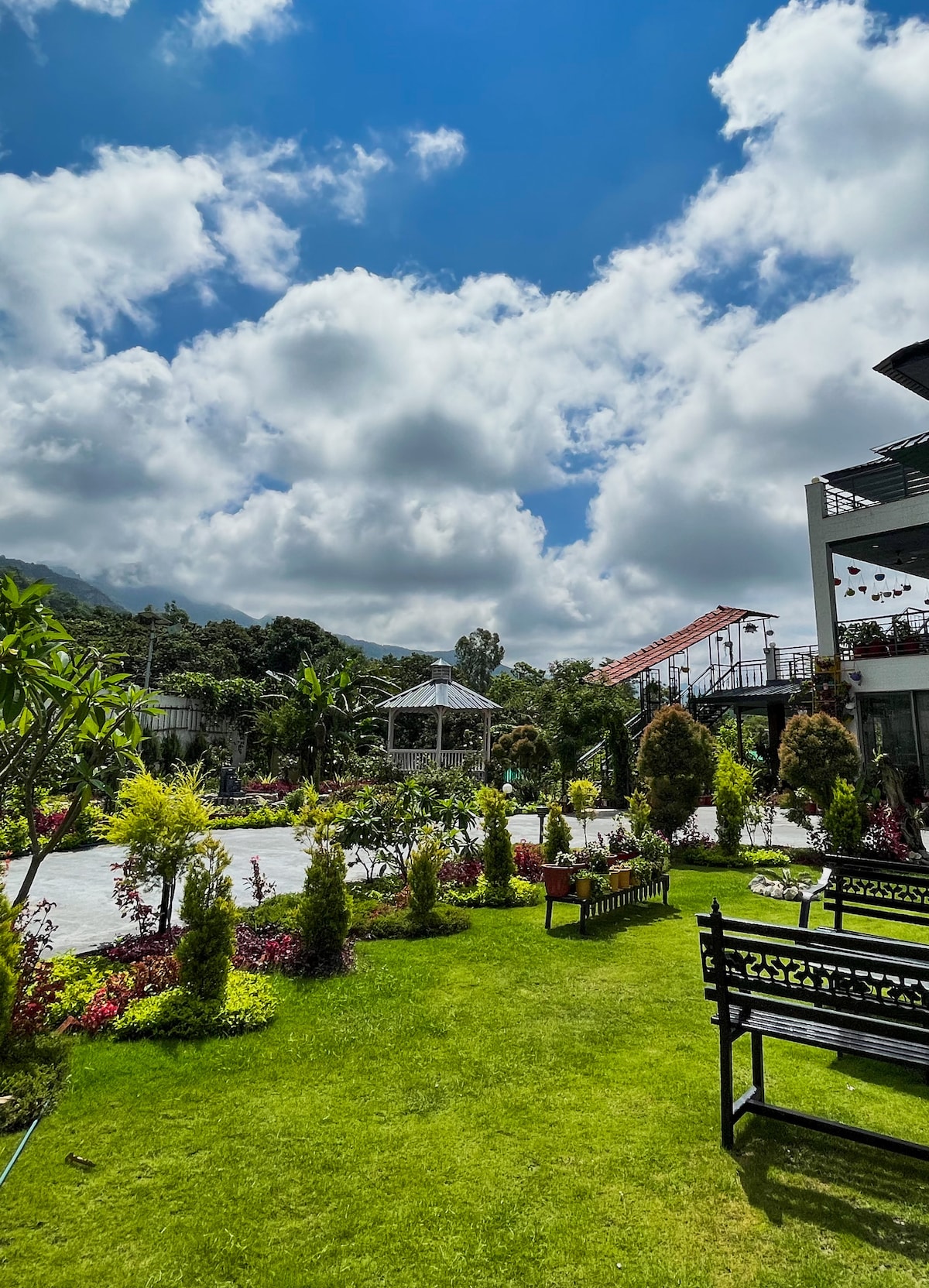 A landscaped garden area is visible, featuring neatly arranged flower beds and greenery. Comfortable benches are positioned for seating, and a gazebo is seen in the background. The sky is partly cloudy, allowing natural light to illuminate the space.