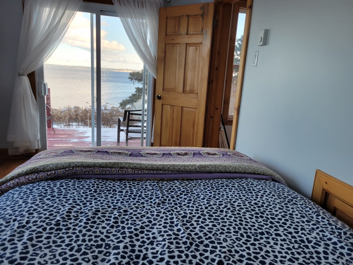A spacious bedroom features a patterned blanket laid across a large bed. Light drapes softly frame the sliding glass door, which opens to a serene waterfront view. A chair stands nearby, complementing the warm wooden accents throughout the room.