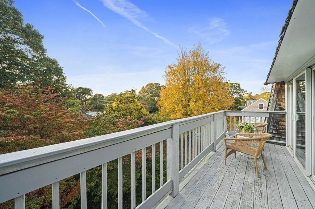 A spacious balcony extends from the main bedroom, featuring a wicker chair positioned to enjoy the view. Surrounding trees exhibit autumn colors, enhancing the natural ambiance. The wooden railings and flooring complement the serene outdoor setting.