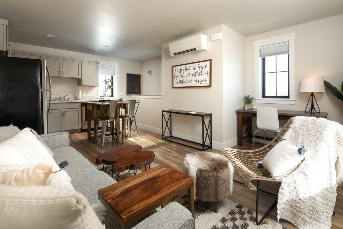 The living area features a blend of modern and rustic elements, including wooden furniture and a cozy neutral palette. A comfortable sofa is positioned next to a stylish chair. A dining table with stools is set next to the open kitchen, creating a versatile space for relaxation and dining.