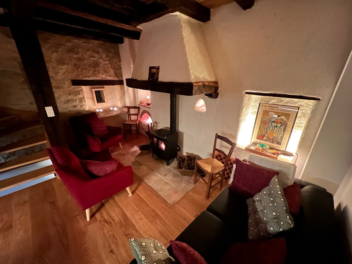 A cozy living area is highlighted by a wood-burning stove, with red upholstered chairs positioned nearby. A black sofa adorned with patterned cushions completes the setting. Soft lighting from wall sconces creates a warm ambiance, complemented by natural wood beams and stone walls.