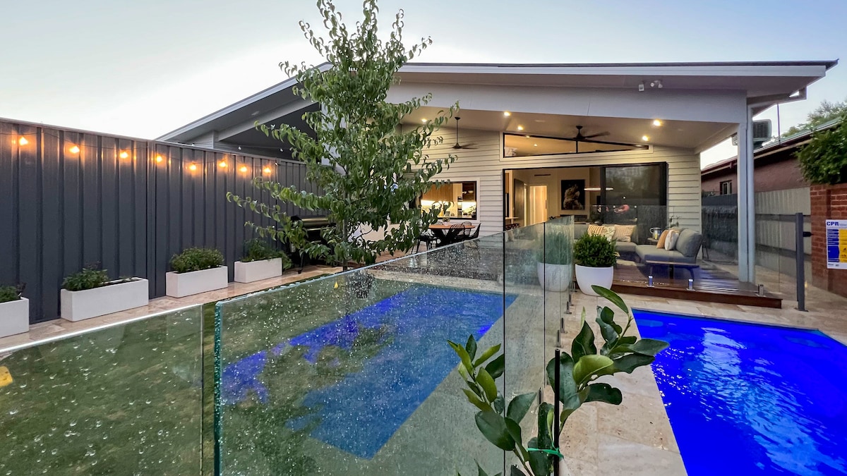 A spacious outdoor area is featured, showcasing a heated pool surrounded by glass fencing. The contemporary home, with large windows, offers ample space for relaxation. Lush greenery in planters enhances the inviting atmosphere, reflecting a blend of indoor and outdoor living.