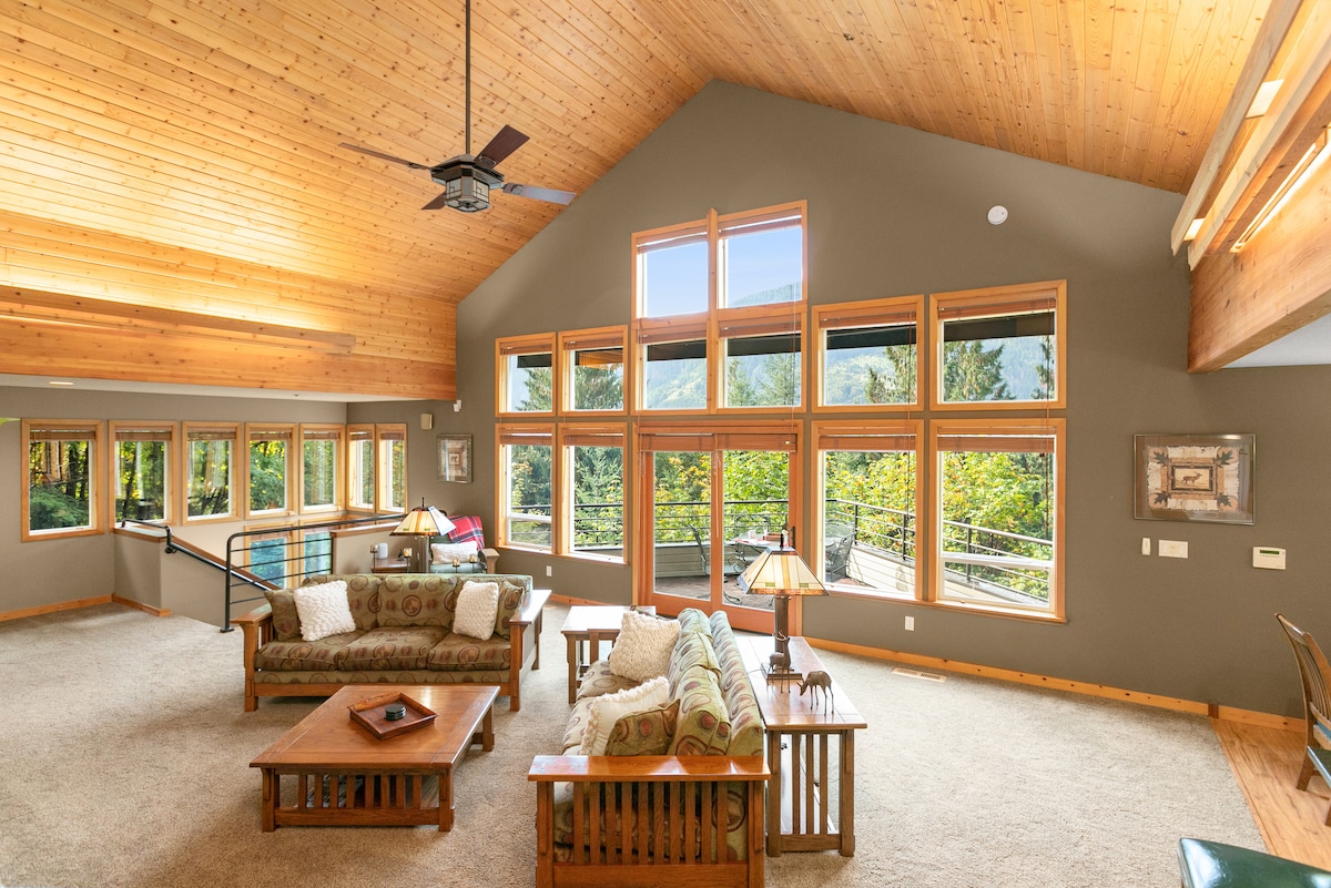 A spacious great room features large windows that provide ample natural light and views of the surrounding trees. A cozy seating arrangement is placed around a wooden coffee table, complemented by a floor lamp. The vaulted wood ceiling enhances the room's airy atmosphere.