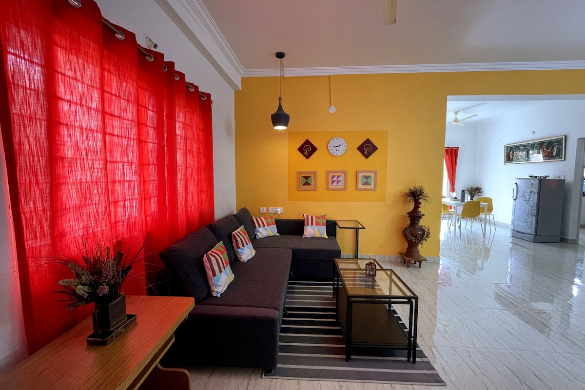 A spacious living area features a dark sofa adorned with colorful cushions, complemented by a striped rug. Bright red curtains frame the window, while decorative wall art enhances the cheerful ambience. A coffee table and side table complete the layout, creating a welcoming space.