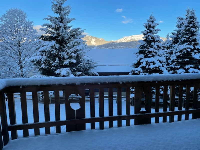 A view from a wooden balcony showcases a snowy landscape, with evergreen trees blanketed in snow. Mountains are visible in the distance under a clear blue sky, creating a serene and wintry scene.