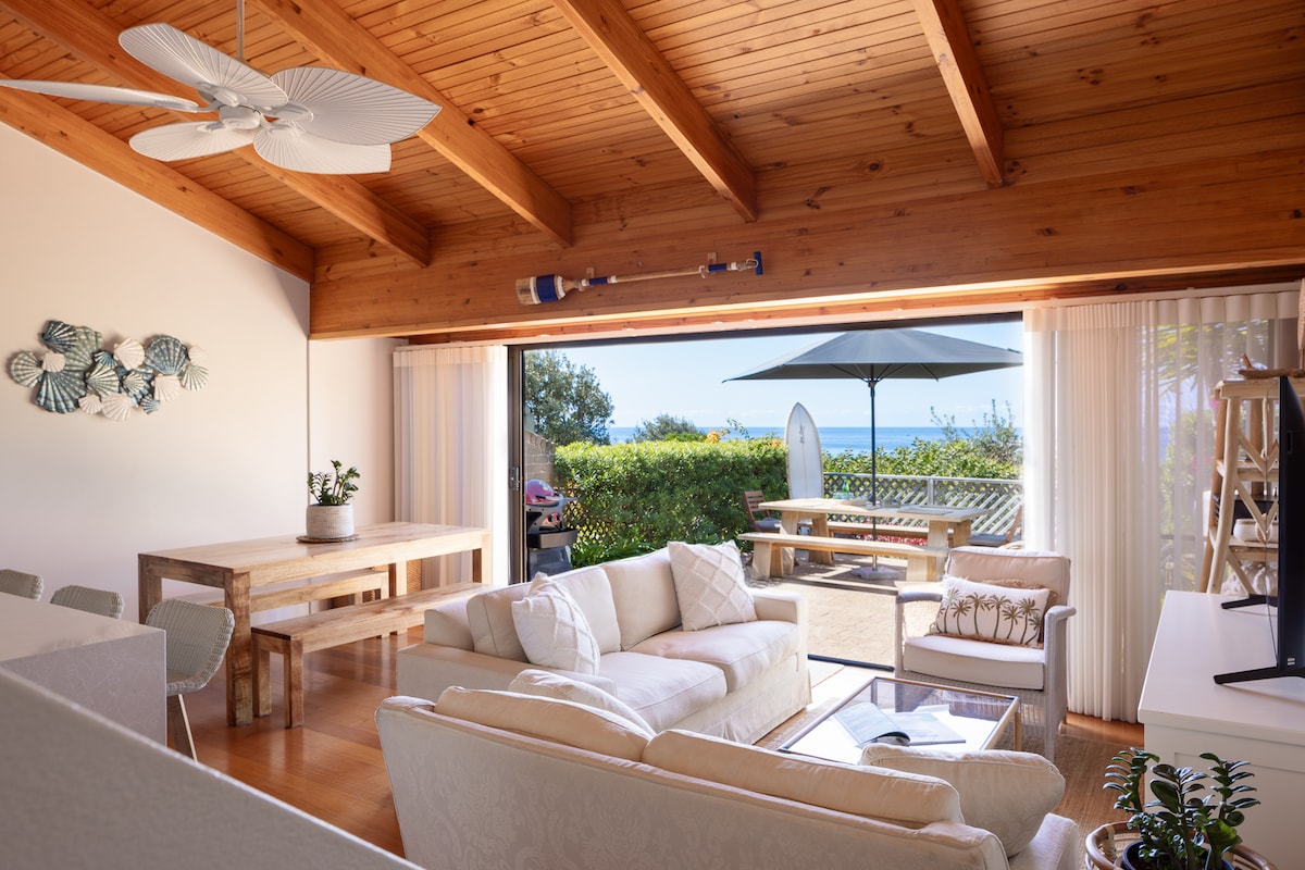 A spacious living area is highlighted by a wooden ceiling and large sliding doors that open to an outdoor space with ocean views. Comfortable seating is arranged around a coffee table, with a dining area featuring a wooden table nearby. Natural light fills the room.