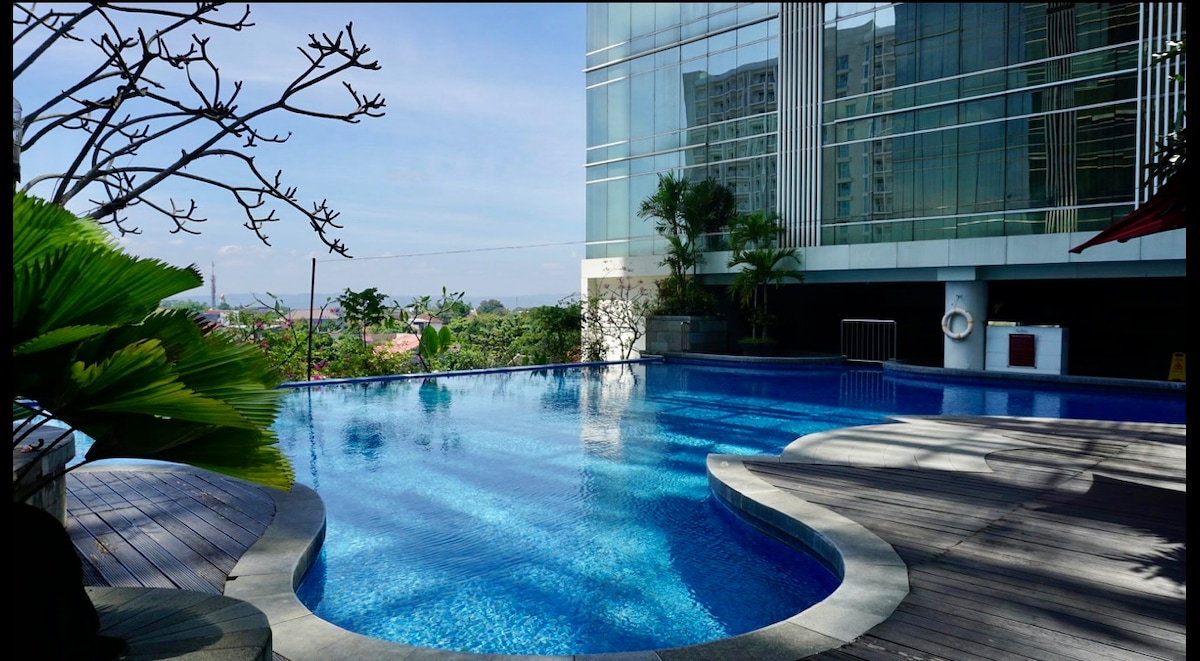 A serene swimming pool is surrounded by lush greenery and a wooden deck. The water gleams under the sunlight, reflecting the modern glass building nearby. Palm trees provide a natural setting, creating a refreshing atmosphere for relaxation.