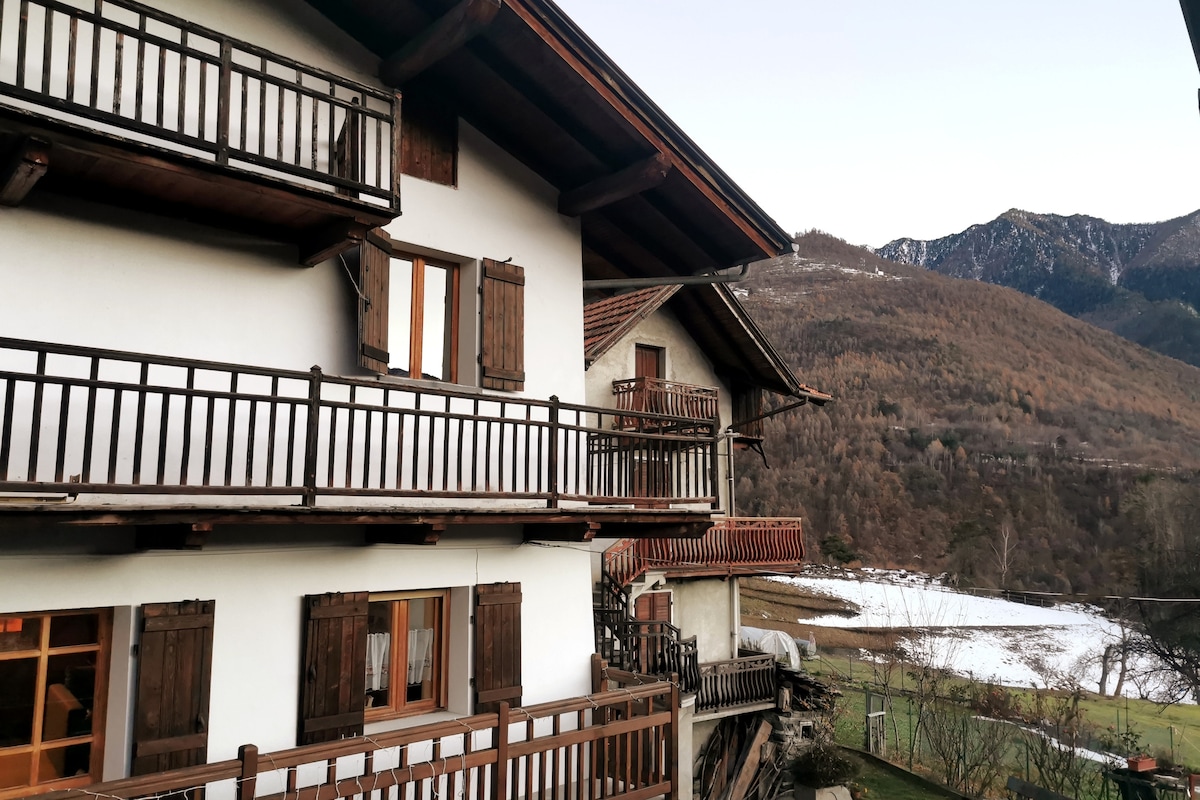 The exterior of the building features multiple wooden balconies with rustic shutters. Surrounding mountains and a snow-covered landscape provide a natural backdrop. The structure blends traditional alpine architecture with inviting outdoor spaces for relaxation.
