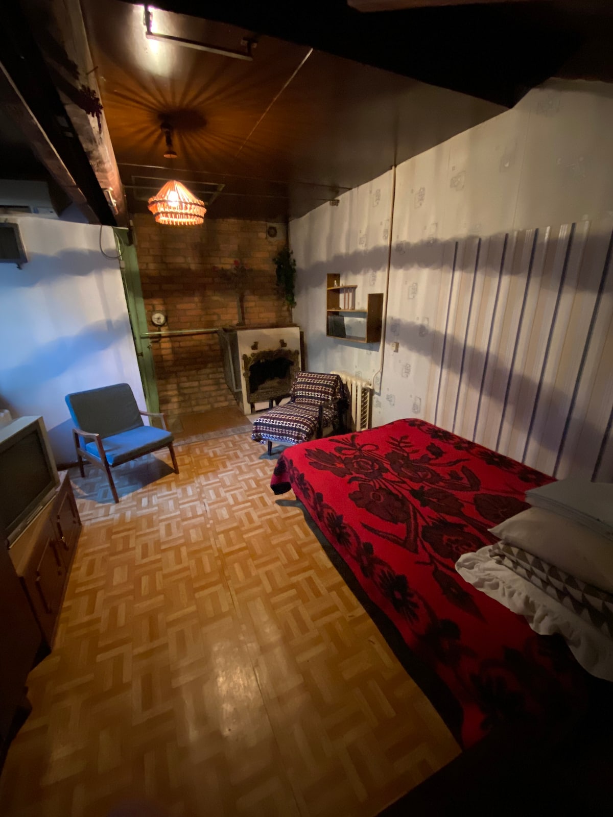 A small room features a spacious bed covered with a red patterned blanket and several pillows. A patterned armchair is positioned nearby, and a vintage television stands on a cabinet against the wall. Exposed brick adds character, complemented by soft lighting from a hanging fixture.