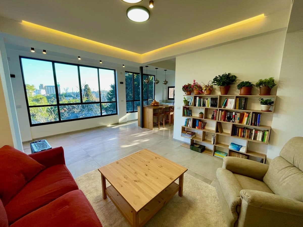 A spacious living area features large windows, offering ample natural light and views of the surroundings. A comfortable red sofa and a beige armchair are arranged around a light wooden coffee table. A bookshelf filled with plants and books adds a touch of greenery and character to the room.