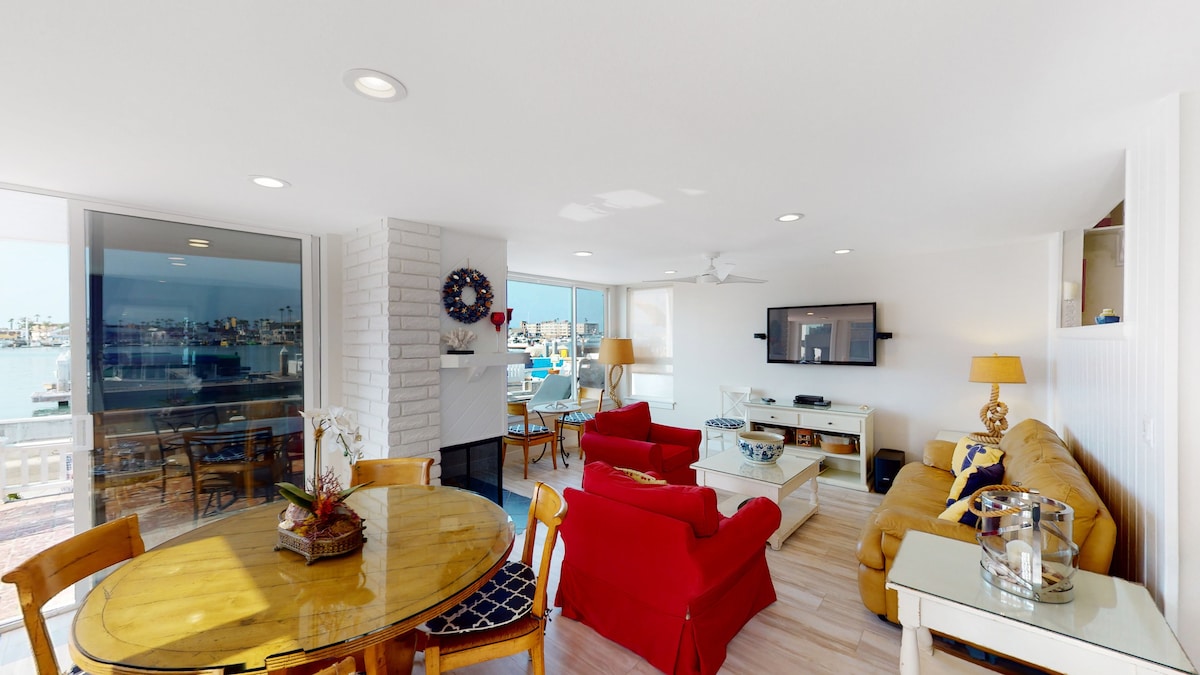 A bright living area features comfortable seating arrangements with a red sofa and armchairs. A dining table is positioned beside a large window, which offers views of the bay. A flat-screen television is mounted on the wall, and natural light fills the space.