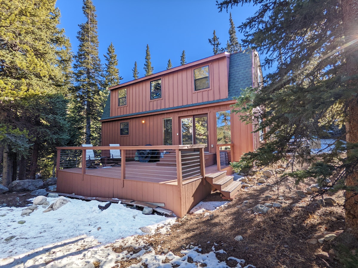 The exterior of the recently renovated mountain home is shown, featuring a modern design in earthy tones. A spacious deck is visible, surrounded by a mix of snow and pine forest. Large windows provide views into the interior, enhancing the connection with nature.