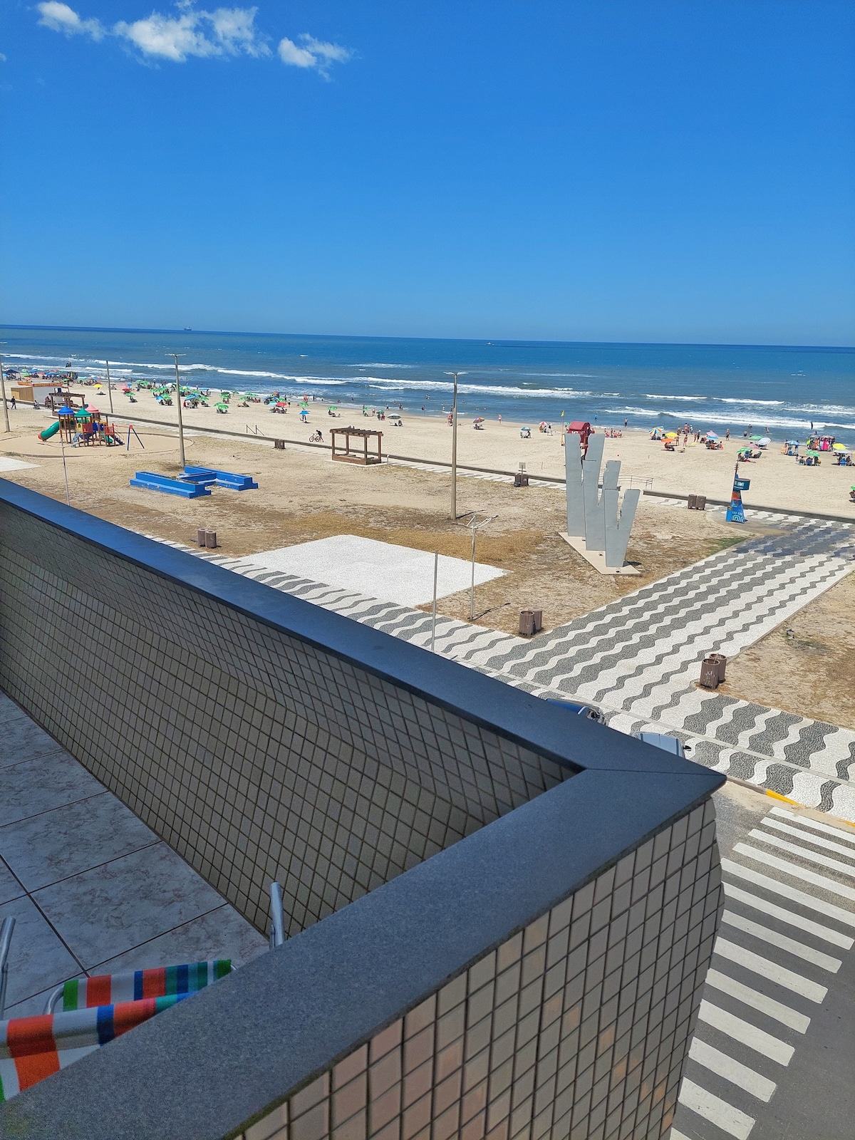 A balcony view reveals a sandy beach and rolling waves under a clear blue sky. Beachgoers are seen enjoying the sun, and colorful umbrellas dot the shore. A patterned walkway leads towards the ocean, highlighting the beachfront's inviting atmosphere.