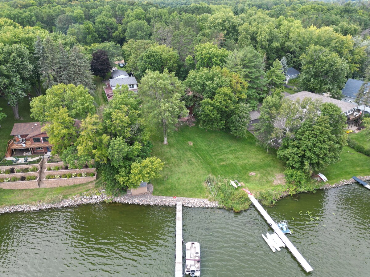 An aerial view captures lush greenery surrounding a spacious lawn, leading down to a calm lake. A private dock is visible, along with a boat moored nearby. The tranquility of the area is evident, framed by trees and residential homes in the background.