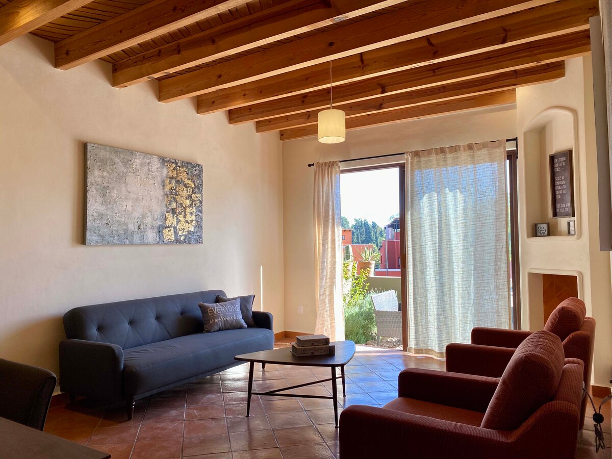 An open living area features a comfortable dark sofa and two cozy red armchairs, arranged around a modern coffee table. Large windows allow natural light to fill the space, with sheer curtains adding a soft touch. Exposed wooden beams highlight the ceiling.