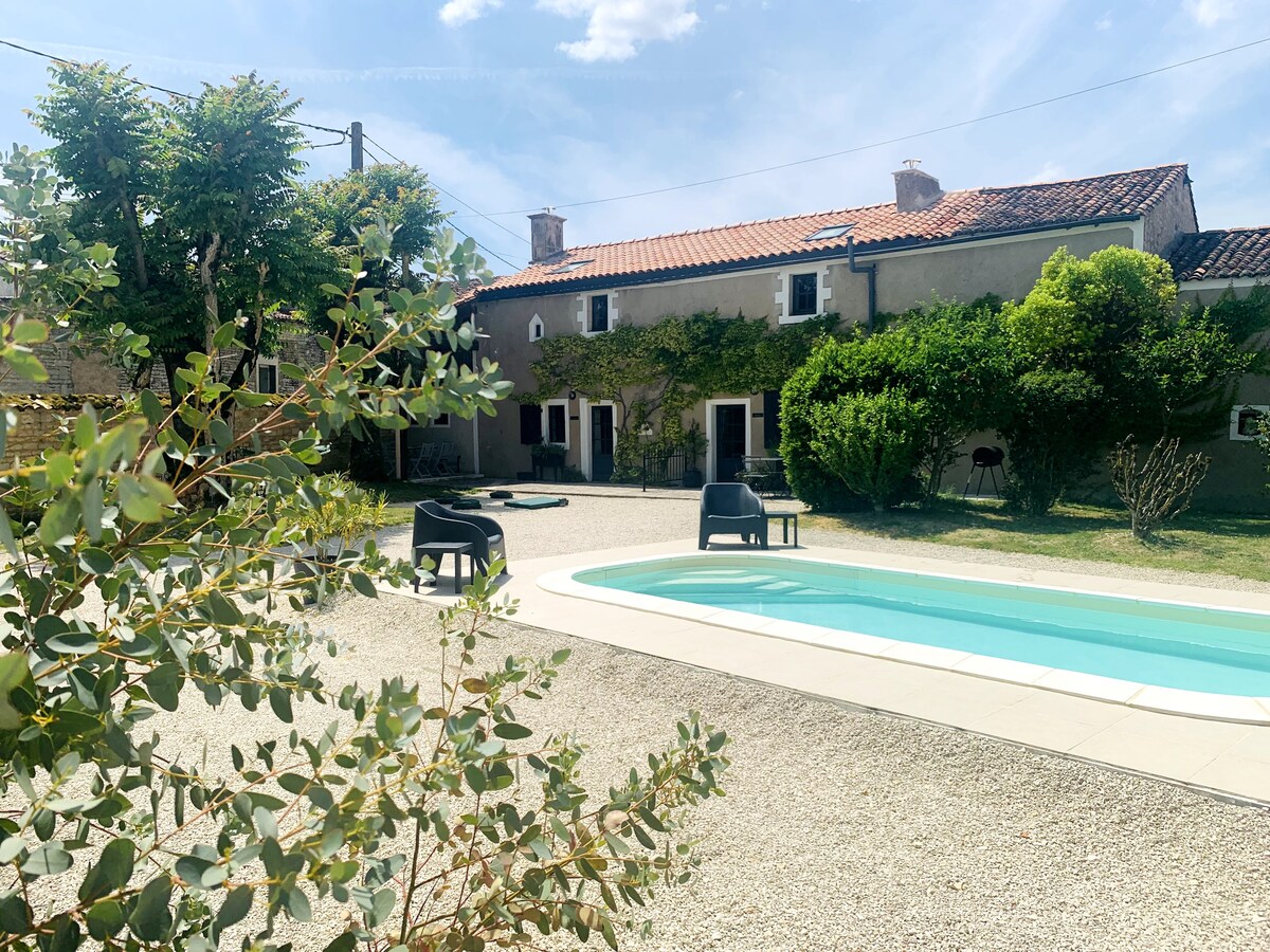 A serene courtyard garden is depicted, featuring a swimming pool surrounded by a gravel area. Comfortable outdoor seating is provided along with lush greenery in the backdrop. The sunlit stone cottage showcases charming windows, completing a peaceful and inviting outdoor space.