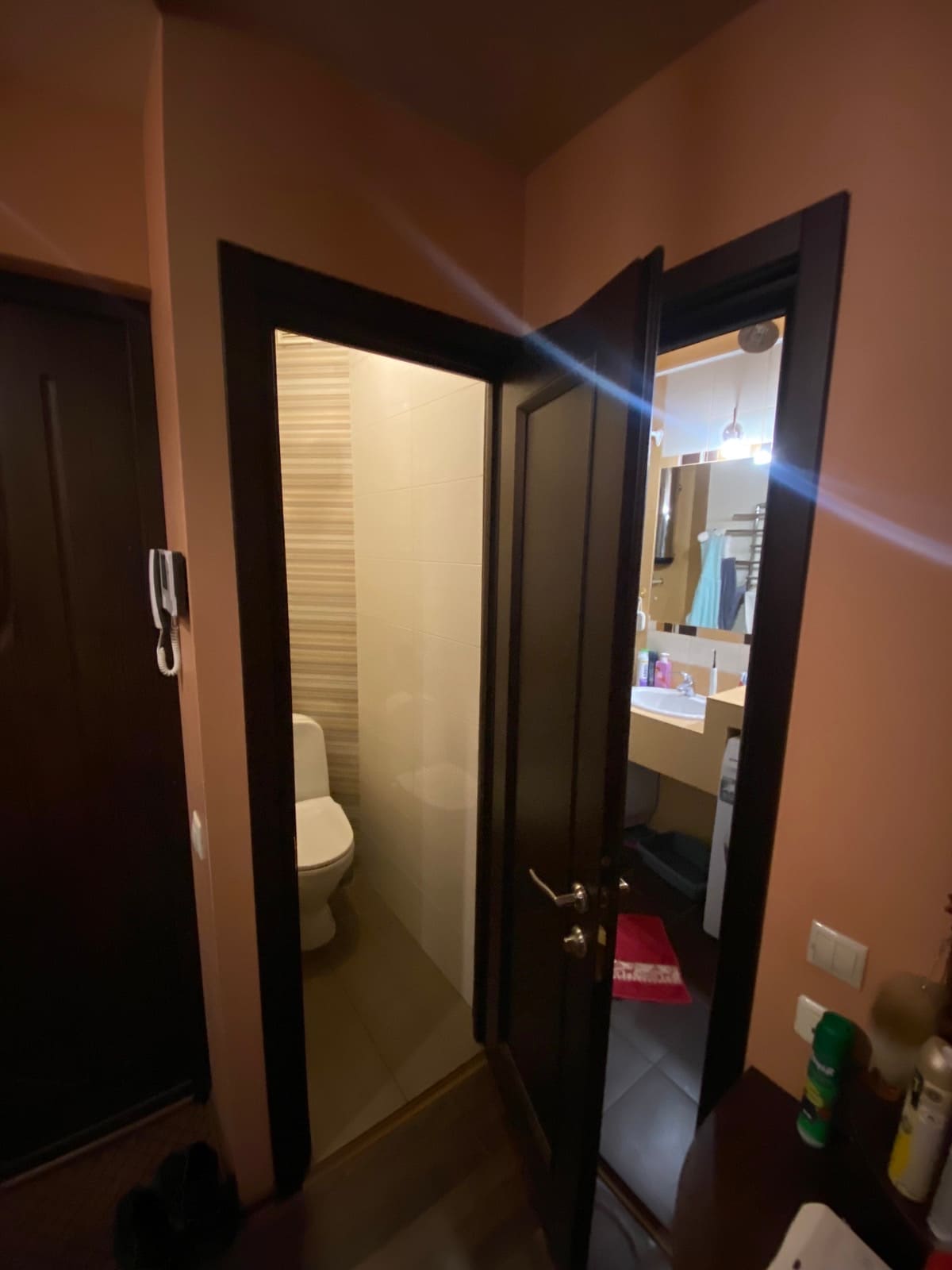 A bathroom doorway is shown, highlighting modern features. The entrance leads to a tidy, compact space with a toilet and a small sink visible. Soft lighting illuminates the area, creating a warm ambiance within the neutral-colored walls.