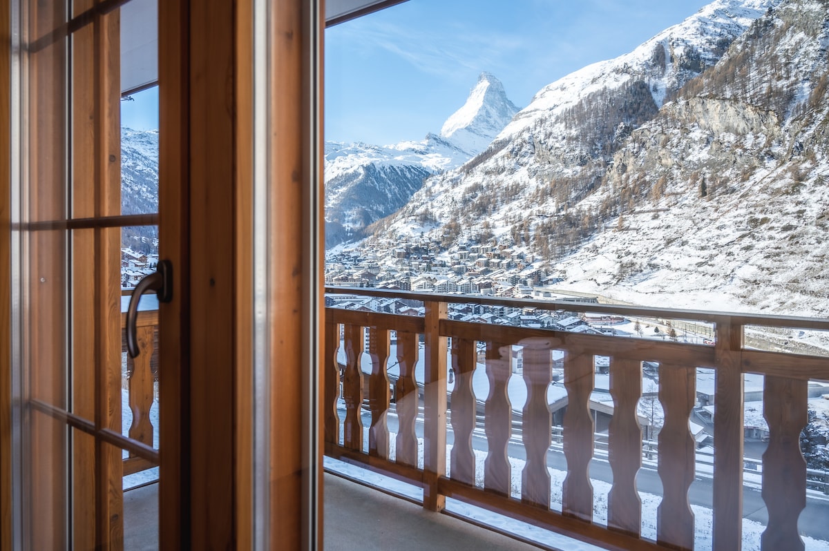 A balcony view showcases snow-covered mountains and valleys under a clear blue sky. The natural wooden railing complements the alpine scenery, framed by large glass doors leading into the apartment. The majestic peaks in the distance create a serene backdrop.