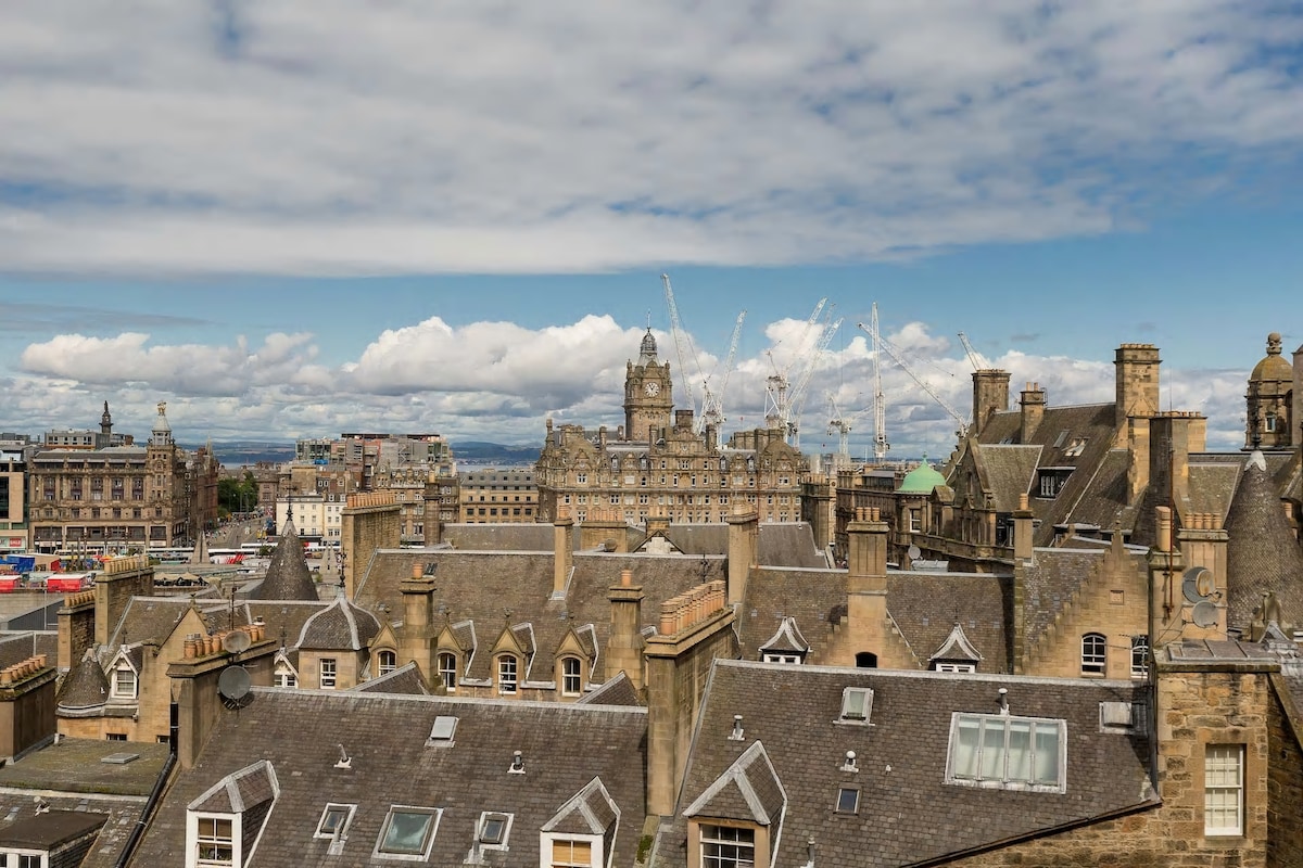 A panoramic view of Edinburgh's cityscape is displayed, featuring a mixture of medieval and Georgian architecture. Rooftops with varying textures and colors can be seen, along with distant outlines of the famous Balmoral Hotel and construction cranes against a backdrop of clouds and blue sky.