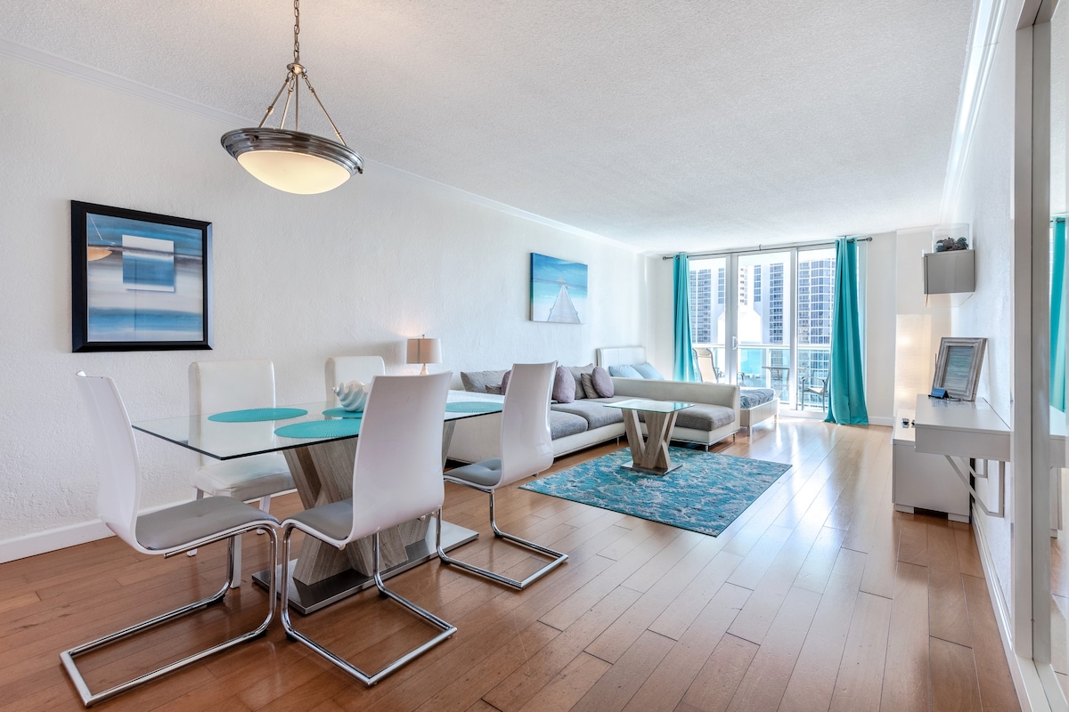 The living area features a bright, open layout with large windows offering views of the city. A modern dining table with clear chairs is complemented by a cozy sectional sofa and a decorative area rug. Light blue curtains add a pop of color to the neutral palette.