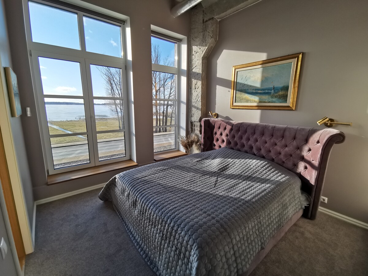 A well-lit bedroom features a spacious tufted bed with a gray quilt. Large windows allow natural light to fill the space, providing views of the outdoors. A decorative painting hangs above the bed, enhancing the room's aesthetic. Soft carpeting covers the floor.
