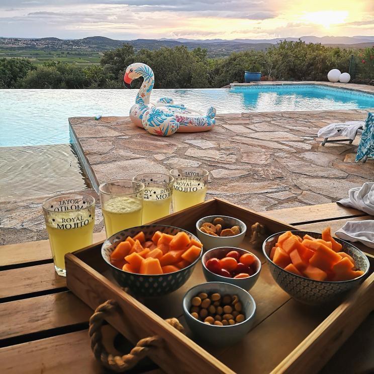 A wooden tray is presented with an assortment of snacks, including cut fruit and nuts. Three glasses filled with light beverages are arranged beside the tray. In the background, a serene infinity pool reflects the sunset, and a colorful pool float adds a playful element.