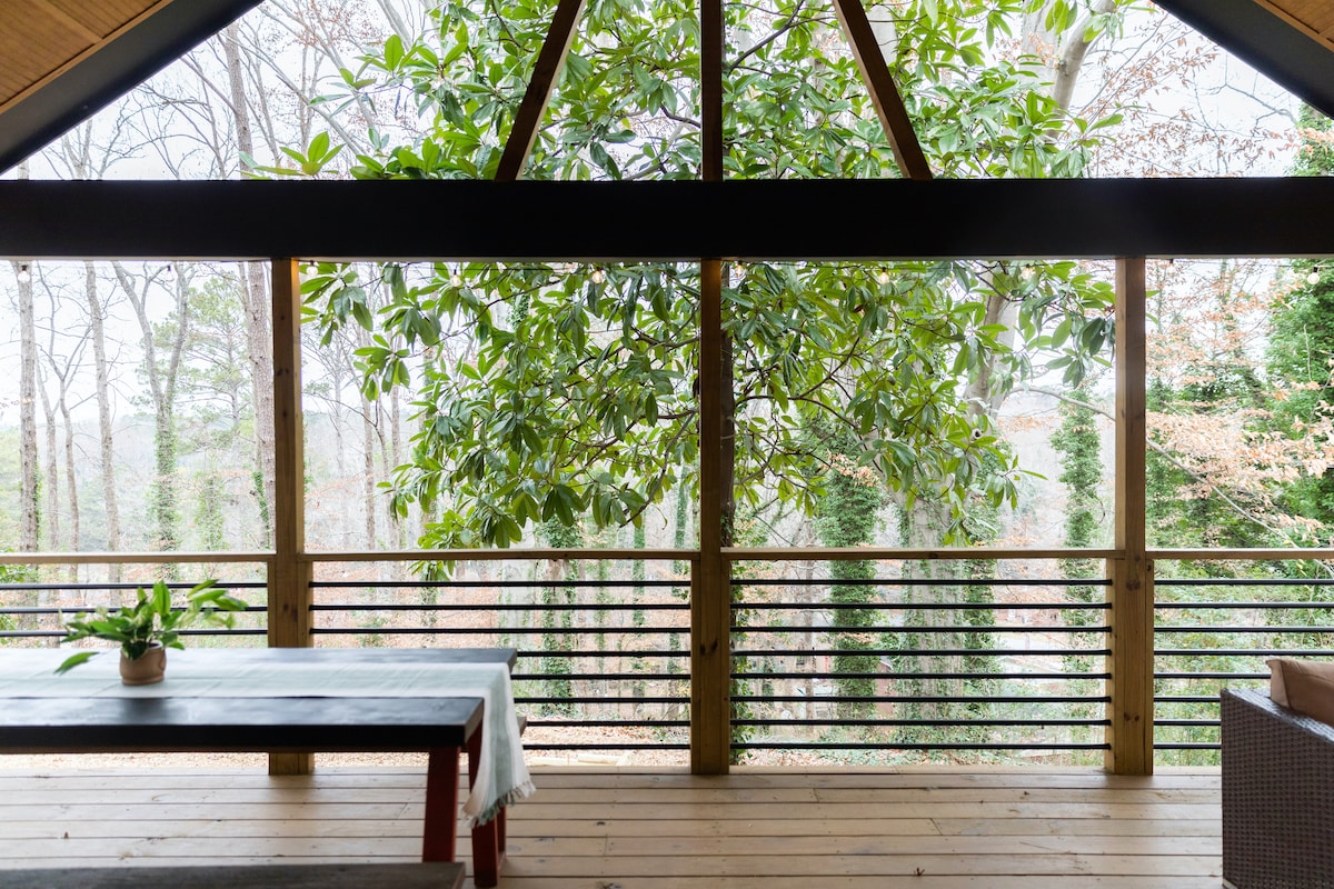 A spacious porch features a wooden floor and an expansive view of the surrounding woods. Large windows allow natural light to fill the area, while a simple table and potted plant invite relaxation in this serene retreat.