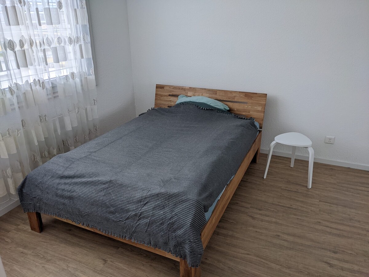 A neatly furnished bedroom features a wooden bed with a gray blanket draped over it. A small side table is positioned beside the bed. Soft natural light is diffused through sheer, patterned curtains, illuminating the light wood flooring.