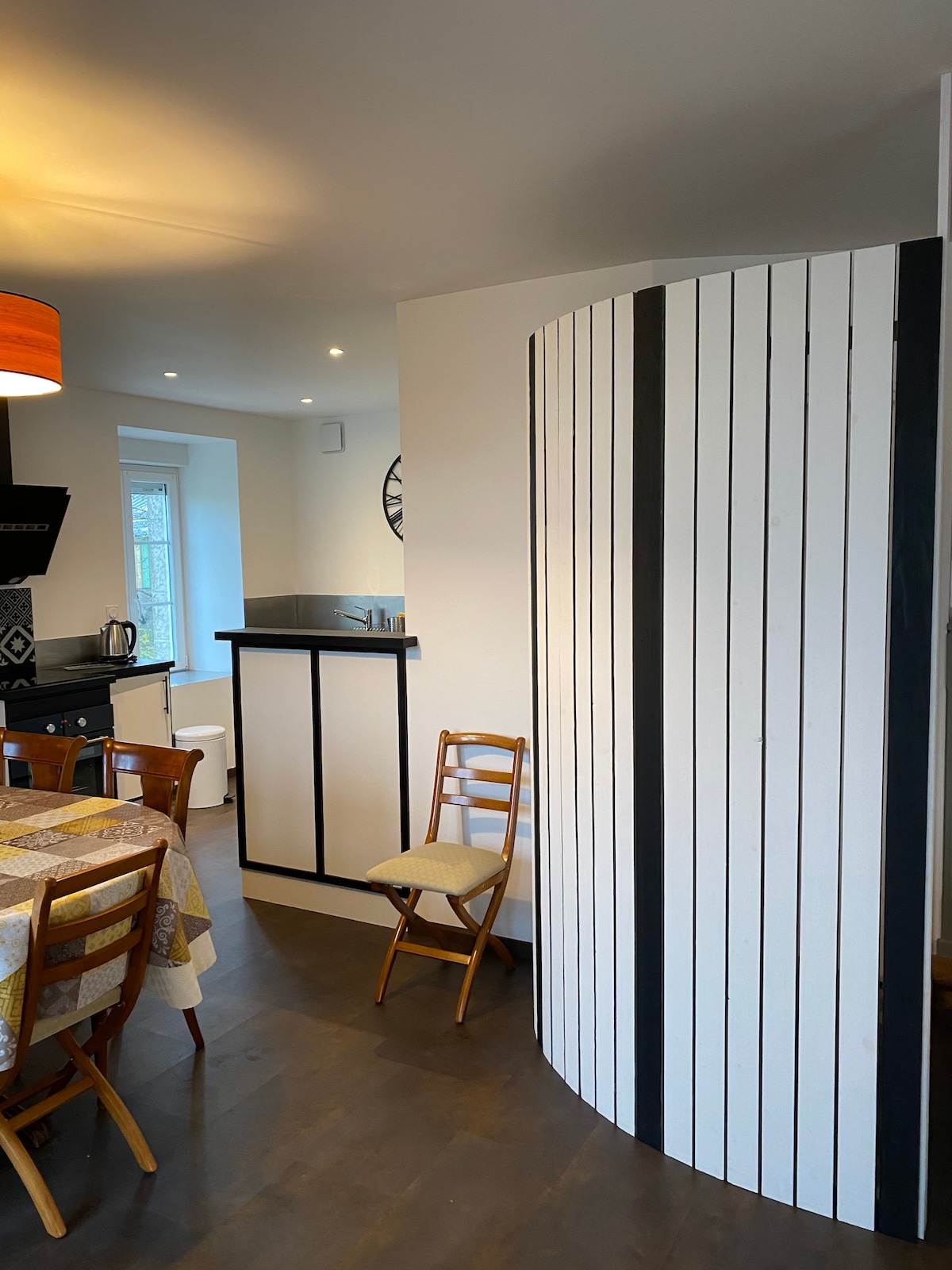 A modern kitchen area features a curved partition with black and white paneling, dividing the cooking space from the dining area. A wooden table with patterned tablecloth is surrounded by four chairs, and a small counter is visible with kitchen essentials.