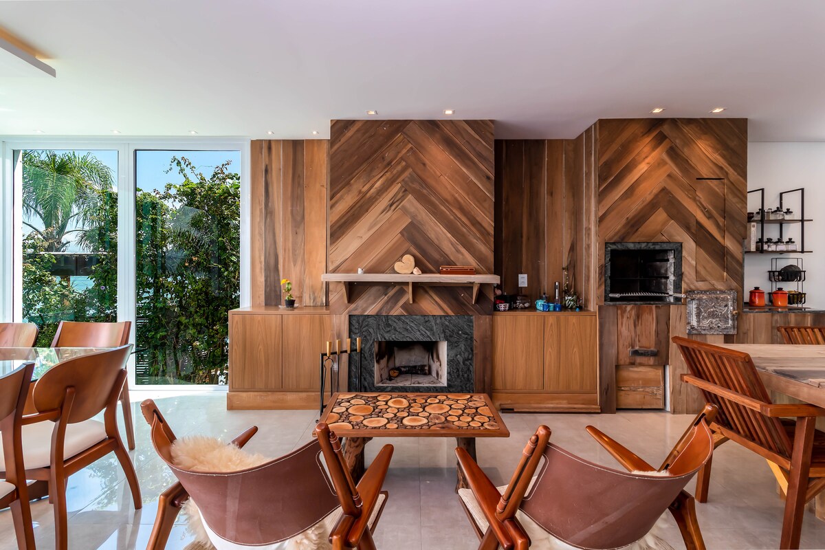 A spacious living area features a modern fireplace with a striking wood panel backdrop. Comfortable seating options include several brown leather chairs surrounding a central coffee table. Large glass windows provide natural light and views of greenery outside, enhancing the open atmosphere.