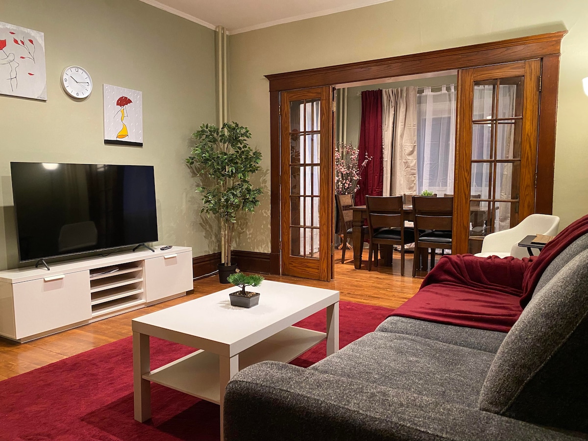 A cozy living area is depicted with a grey sofa accented by a red throw. A flat-screen TV is positioned on a light-colored media console. A coffee table with a small plant sits in front, while a dining area is visible through the French doors, featuring a wooden table and chairs.