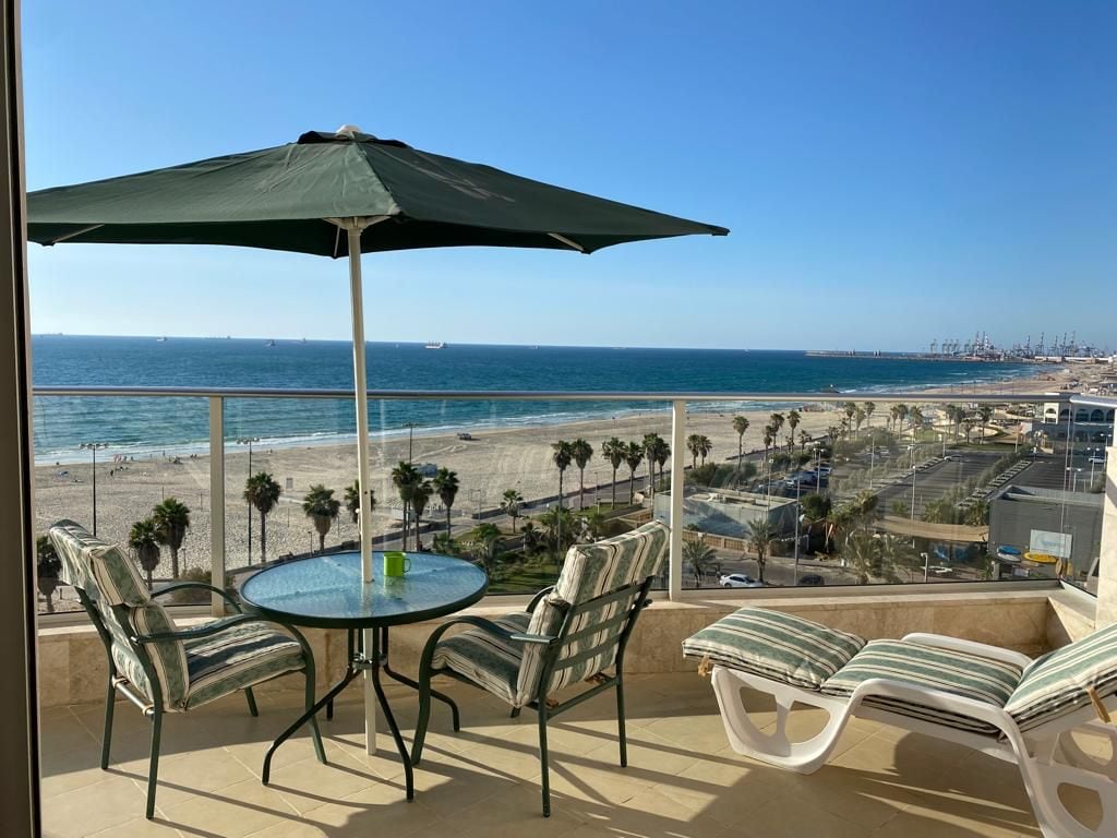 A spacious balcony features a round glass table accompanied by two chairs and a lounge chair, all adorned with green and white cushions. An umbrella provides shade, offering an unobstructed view of the sandy beach and the ocean, with palm trees framing the scenery.